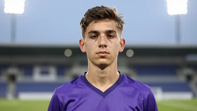 A 20-year-old left-back in a solid purple football kit, standing on a football pitch with blurred stadium lights in the...