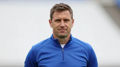 A 40-year-old male goalkeeper coach, with a strong build and short brown hair, stands in front of a white background...