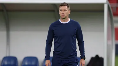 A 25-30 year old, athletic, male football coach, wearing a dark blue, long-sleeved, polyester shirt, with a white collar...