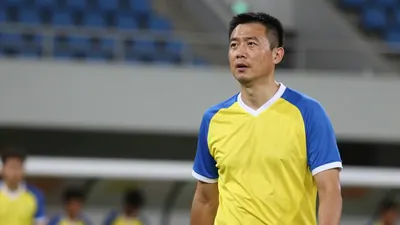 A 35-year-old male assistant coach, wearing a yellow and blue jersey with a white collar, stands on the touchline...