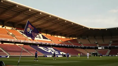 A tense pre-match scene on a rain-glossed Ligue 1 pitch, two anonymous players - one in a muted violet jersey, the other...