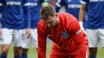A Hamburg SV goalkeeper in a red jersey, mid-30s, frustrated after a misplaced pass, head down, surrounded by Everton...