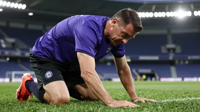 A Toulouse FC groundskeeper in a purple and black kit, mid-30s, muscular build, kneeling on the Stadium pitch, inspecting...